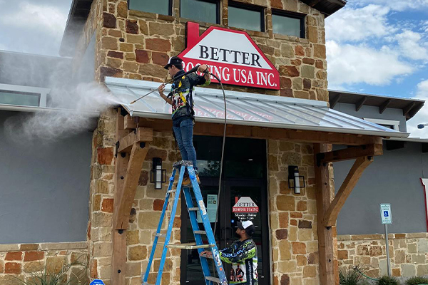 A BioCleaners USA employee on a ladder cleans a ceiling at the entrance of a commercial building, using the softwashing technique.