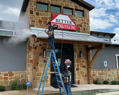 A BioCleaners USA employee on a ladder cleans a ceiling at the entrance of a commercial building, using the softwashing technique.